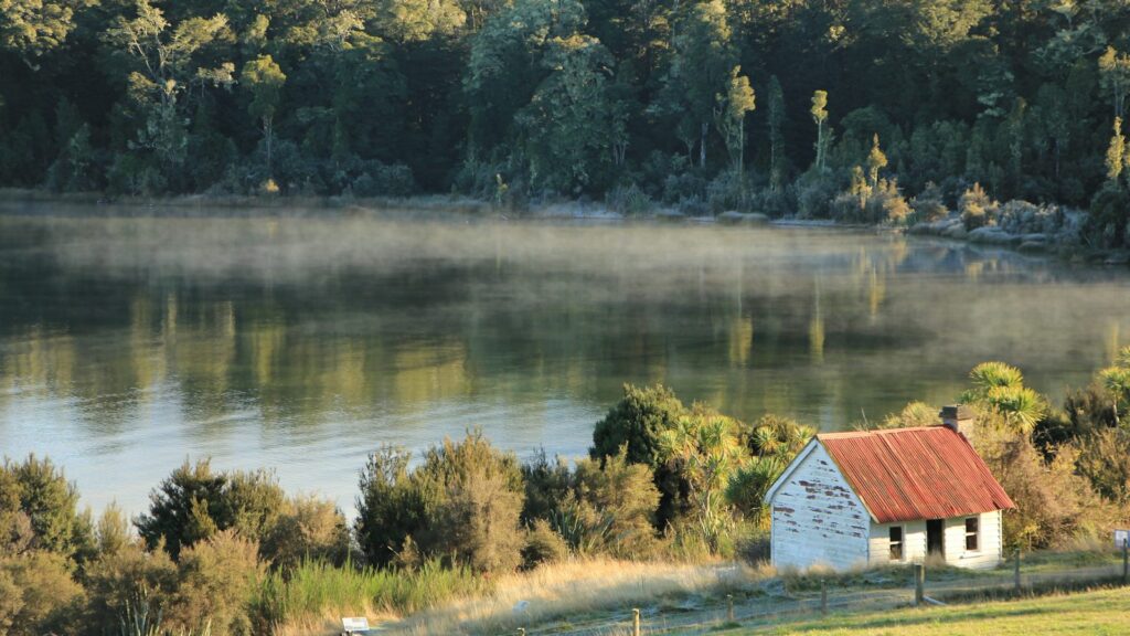 A small white cottage with a red roof beside a lake as morning mist rises off the water below a forested hillside