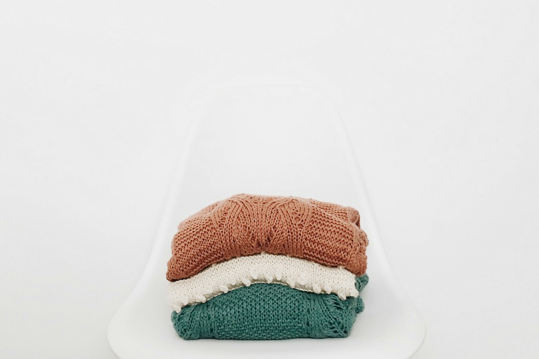 Three folded knit sweaters in terracotta, cream and teal stacked on a white chair