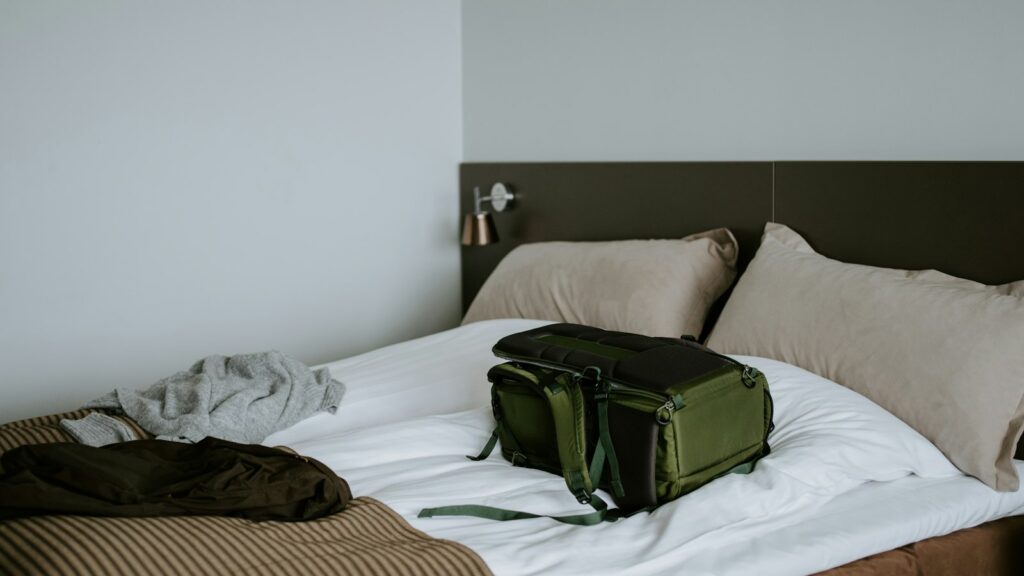 An olive-green backpack resting on a freshly made bed beside a folded striped blanket and a few unpacked clothes
