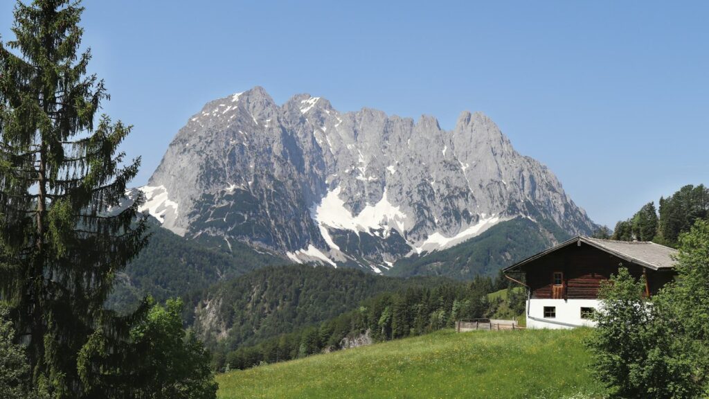 A wooden alpine house on a green meadow below a towering grey limestone massif under a clear sky