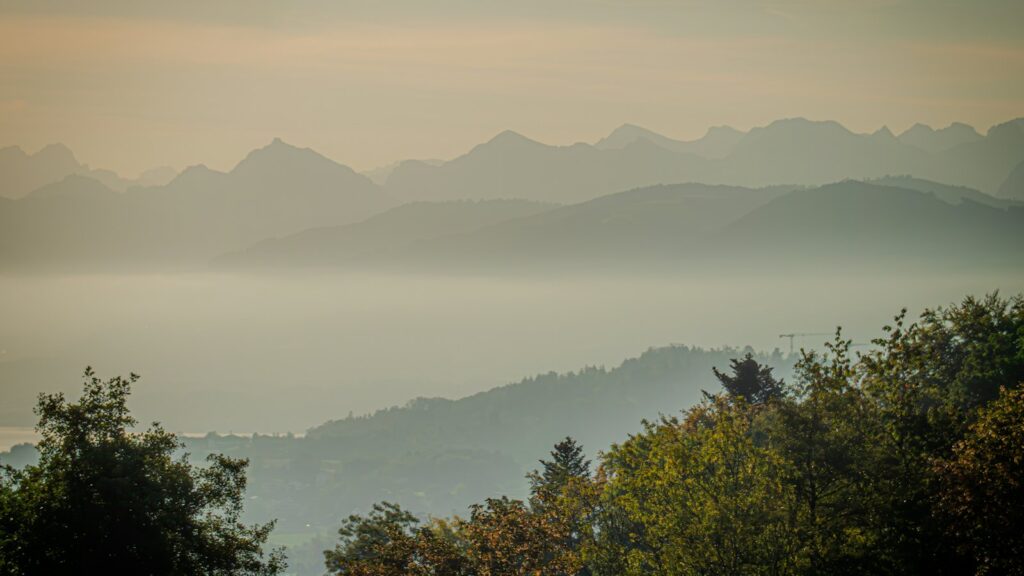 Layered mountain ridges rising above a valley filled with soft morning mist, with autumn trees in the foreground