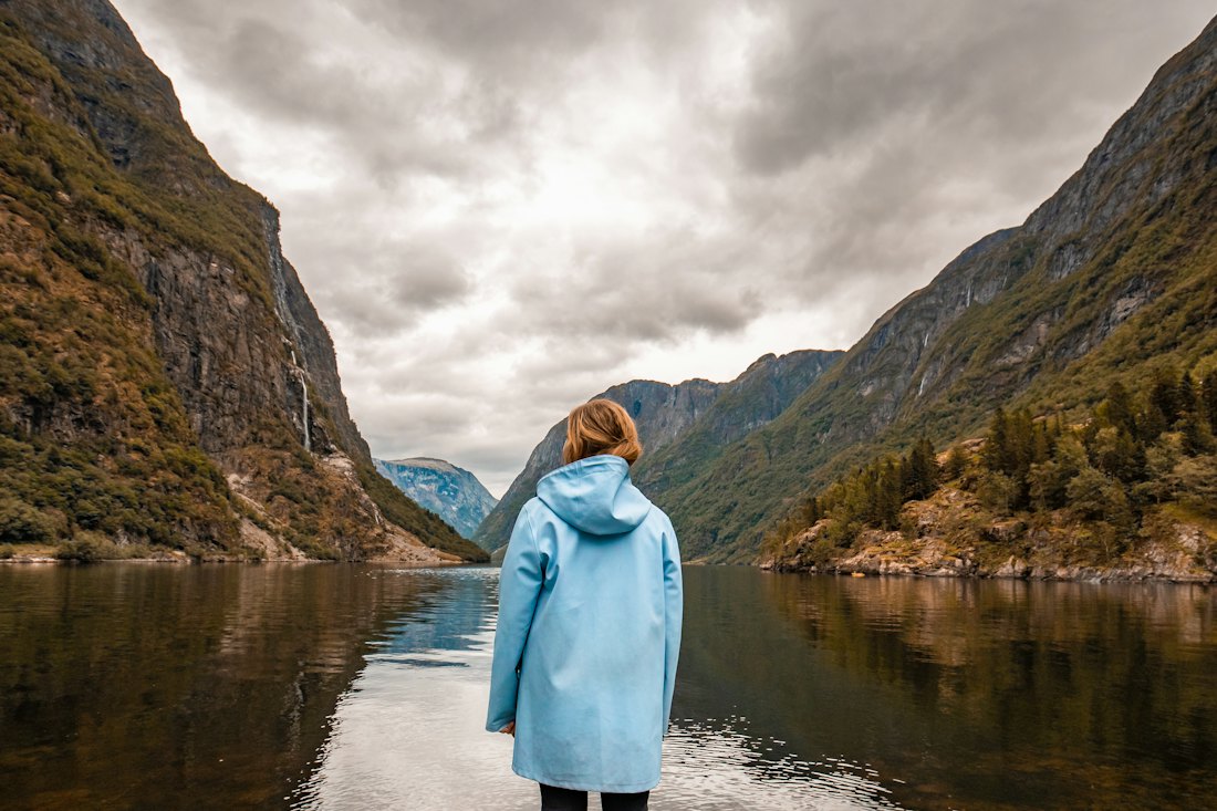 A traveller in a light blue rain jacket standing by a still fjord between steep green mountains