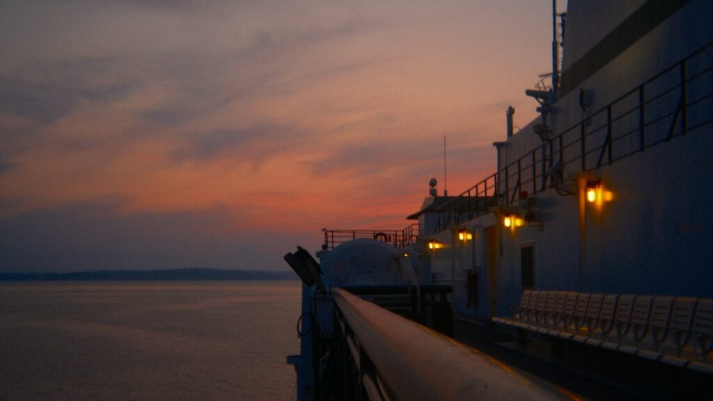 The deck of a ferry at dusk with warm lamps glowing and a pink and amber sky over calm water