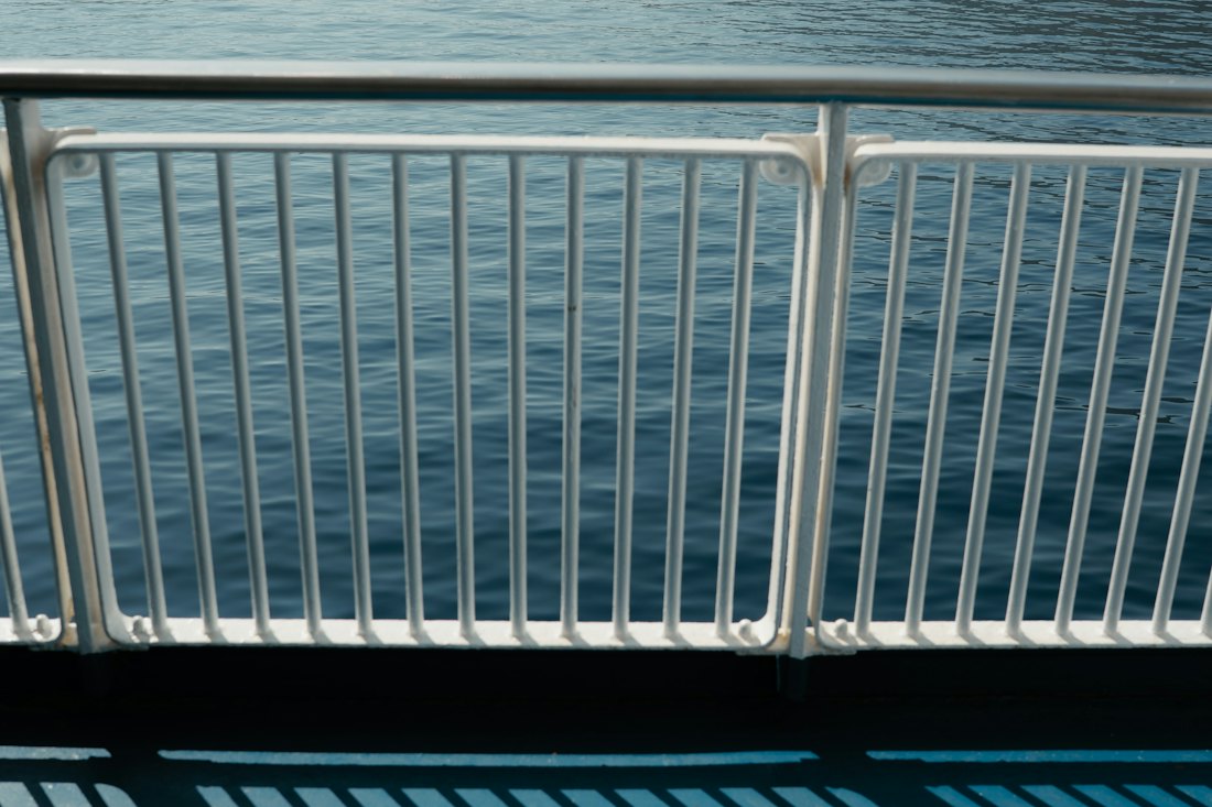 A white ship railing casting striped shadows on a blue deck above calm sea water