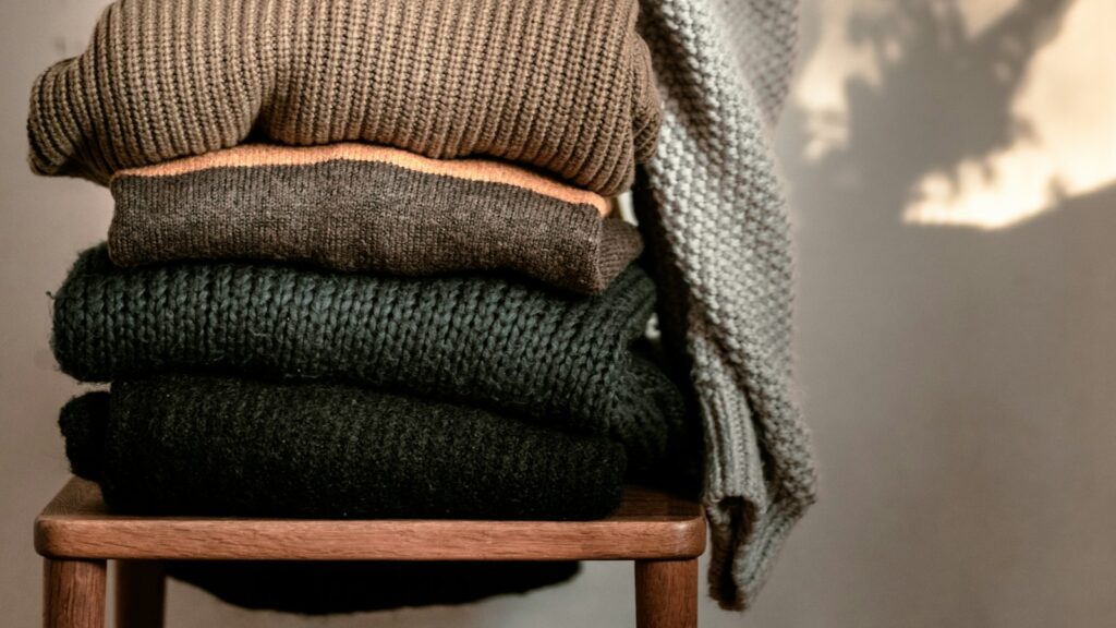 A neat stack of folded wool sweaters in brown, charcoal and forest green on a wooden stool in soft sunlight