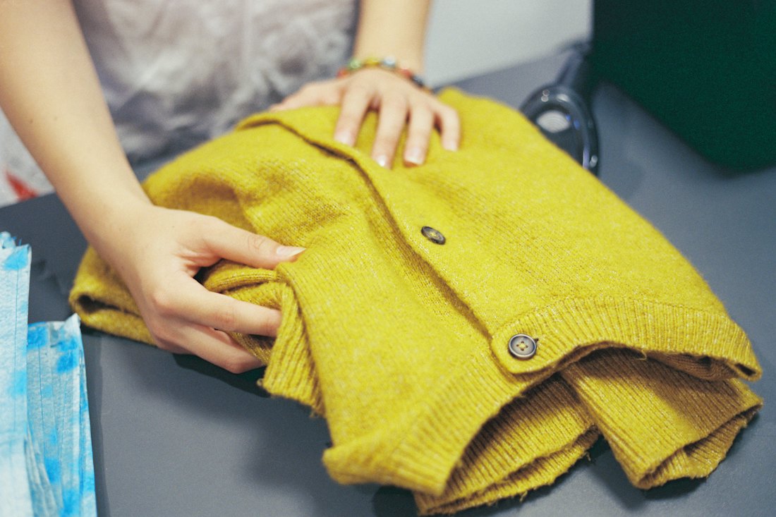 Hands smoothing a folded mustard-yellow cardigan on a grey surface