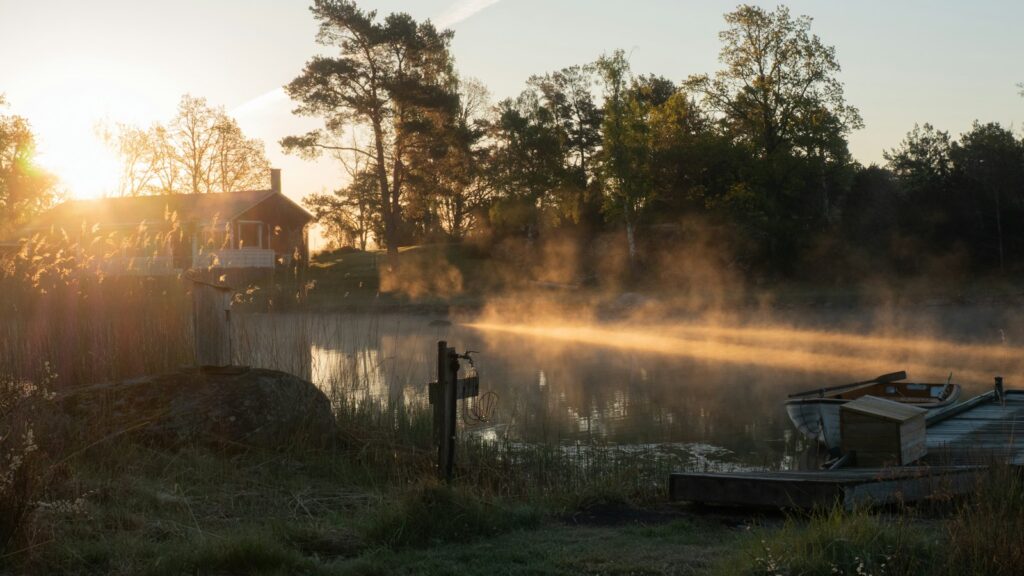 Morning sun rising behind a lakeside house as mist drifts over the still water and a small wooden boat
